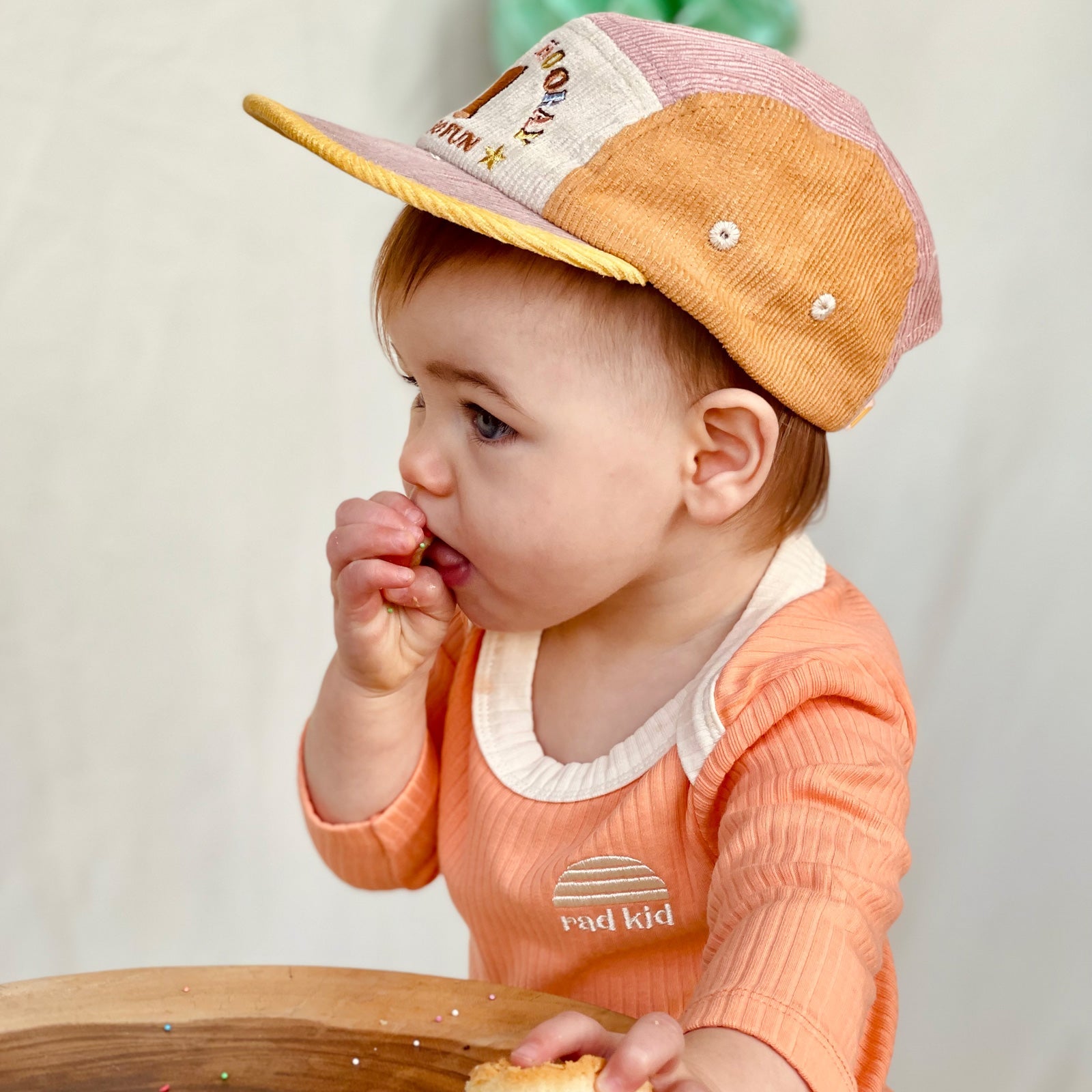 Party time photo, one year old  child wearing Banabae Peach Ribbed Organic Cotton Long Sleeve Onesie with contrast natural neck bind and Rad kid/Banabae Sun Logo embroidered on right hand . Child is also wearing the  1st Birthday Cord Cap musk Spliced, sh