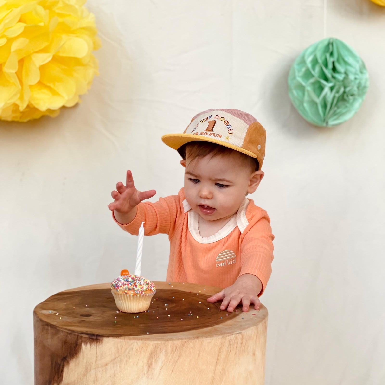 Party time photo, one year old  child wearing Banabae Peach Ribbed Organic Cotton Long Sleeve Onesie with contrast natural neck bind and Rad kid/Banabae Sun Logo embroidered on right hand . Child is also wearing the  1st Birthday Cord Cap musk Spliced, sh