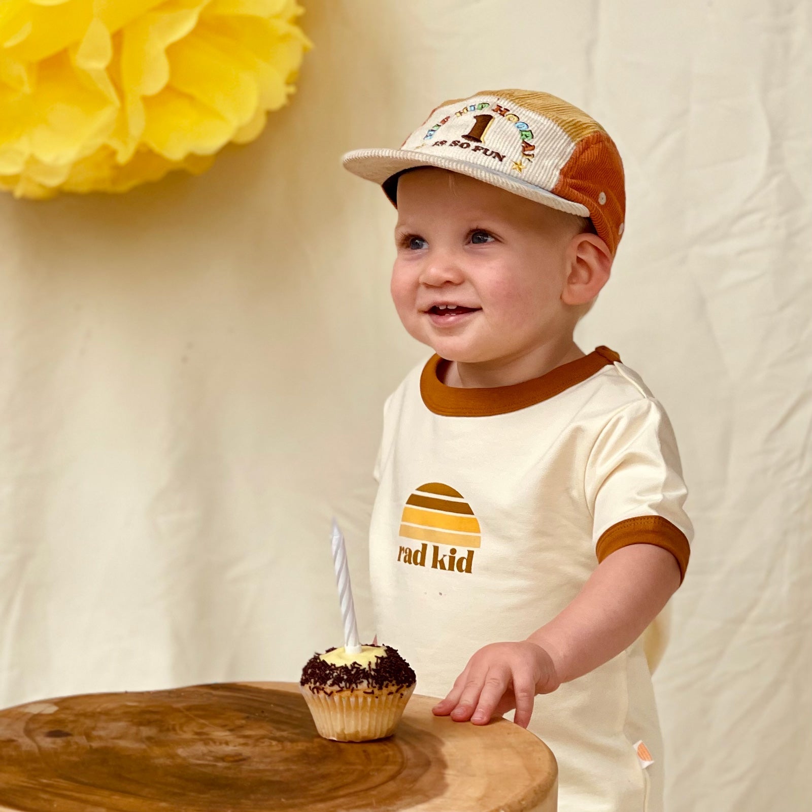 Child wearing 1st Birthday Cord Cap and Rad Kid T-shirt with birthday cupcake in front of them.