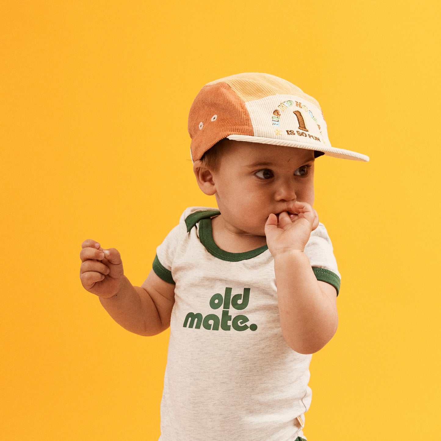 Child wearing the 1st Birthday Cord 5 Paneled Cap.  which is a colour blocked design in blue, orange, natural and yellow . Front of cap is natural with HIP HIP HOORAY 1 IS SO FUN embroidered in multicolored thread. Blue peak with contrast Natural bind.  T