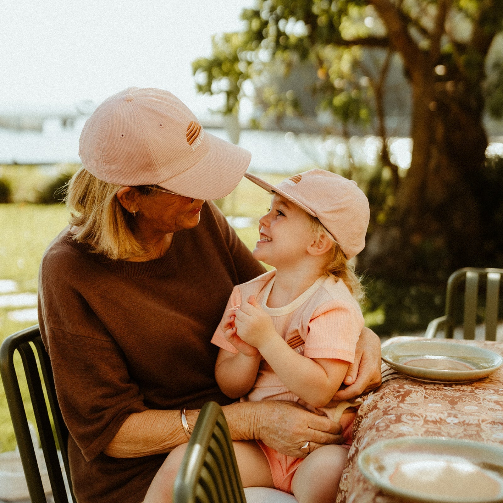 Life style image of grandmother with granddaughter sitting on her lap, both wearing Banabae pink cord caps.