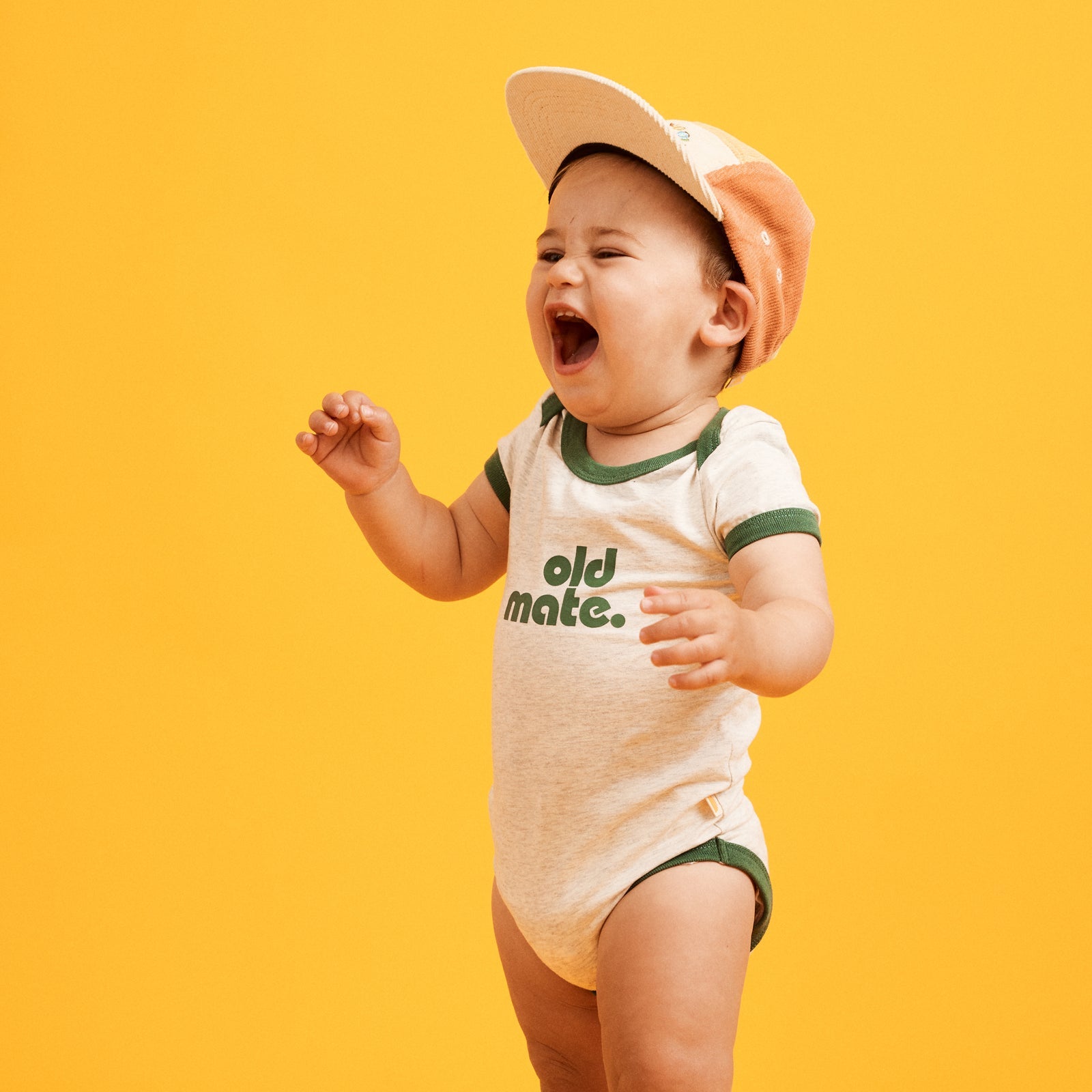 Image of happy one year old wearing a cap and natural grey marled based onesie  with contrast green ringer bind at sleeve, neckline and hemp. Old Mate in green printed at center front.
Snap Button opening at crotch for easy changing and dressing. 