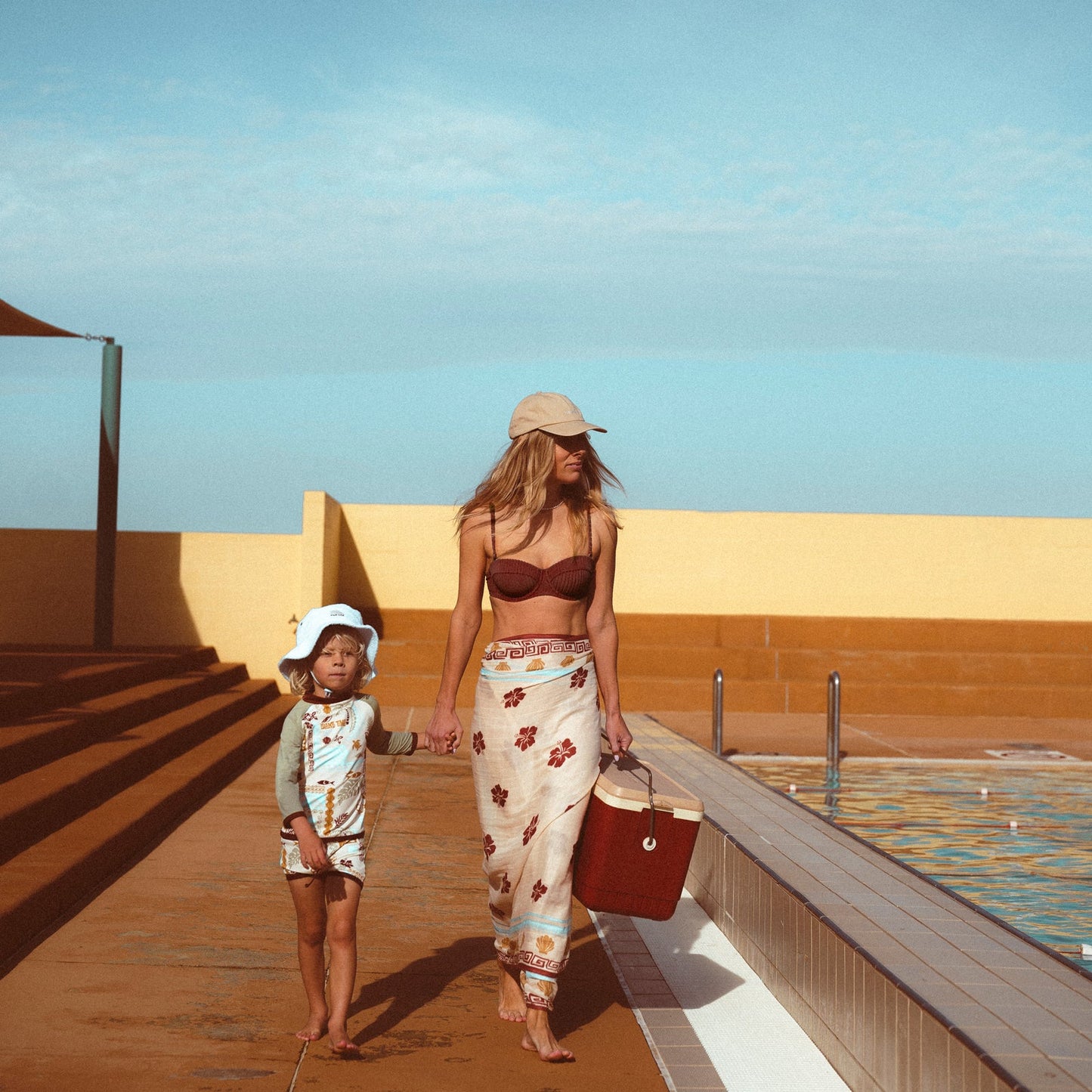 Lifestyle image of mother and son at an open air pool walking together holding hands beside the pool.  The mother is wearing the Banabae washed sand cap in organic cotton twill, Banabae embroidery at the center front in contrast white.  Brown bikini and B