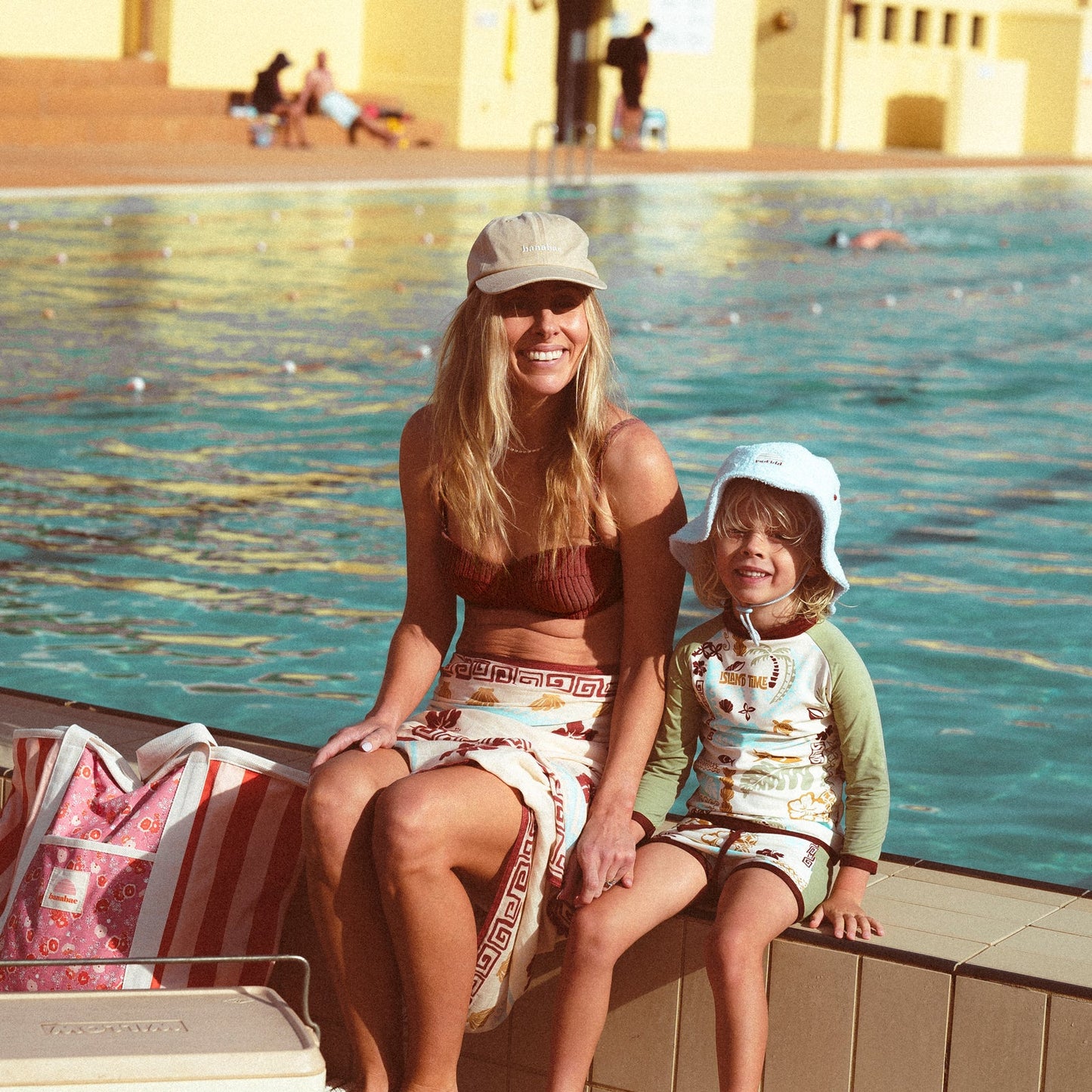 Lifestyle image of mother and son at an open air pool sitting together at the side of the pool.  The mother is wearing the Banabae washed sand cap in organic cotton twill, Banabae embroidery at the center front in contrast white.  Brown bikini and Banabae