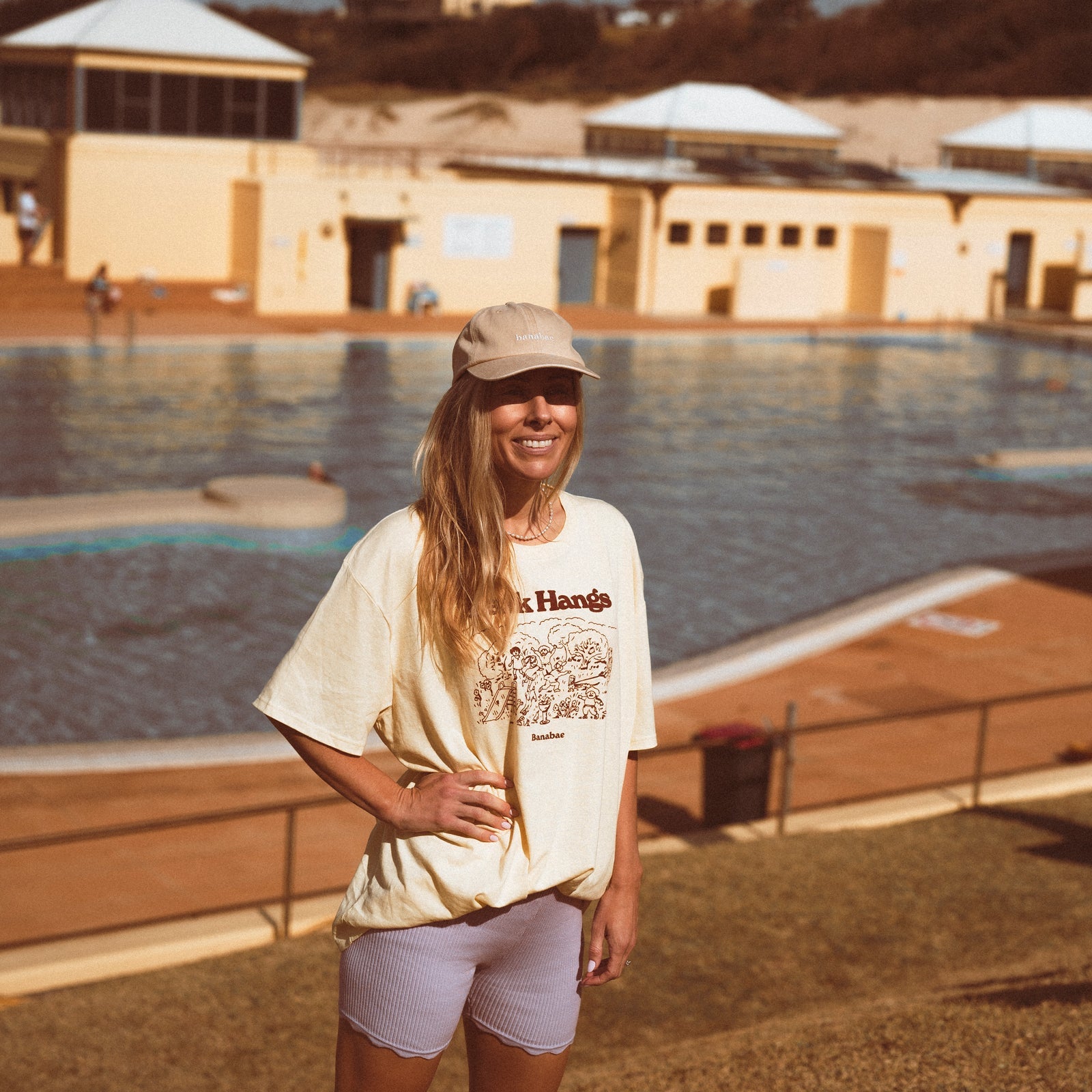 Lifestyle image of Woman wearing  the Banabae washed sand cap in organic cotton twill, Banabae embroidery at the center front in contrast white.  Oversized Park Hangs Tee in yellow with brown print and lilac ribbed bike shorts. Standing in front of a pool