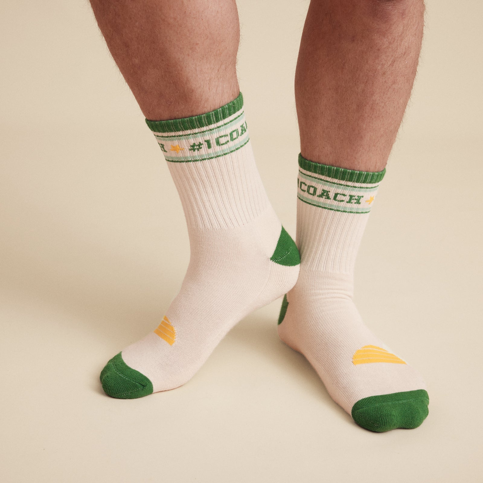 close up Image of socks  worn on the body.   #1 Coach socks, White crew socks with #1 coach in Green written horizontally, bordered with a green and aqua stripe. Toe and heal pads in contrast green with Banabae written along the sole in yellow, and the ic