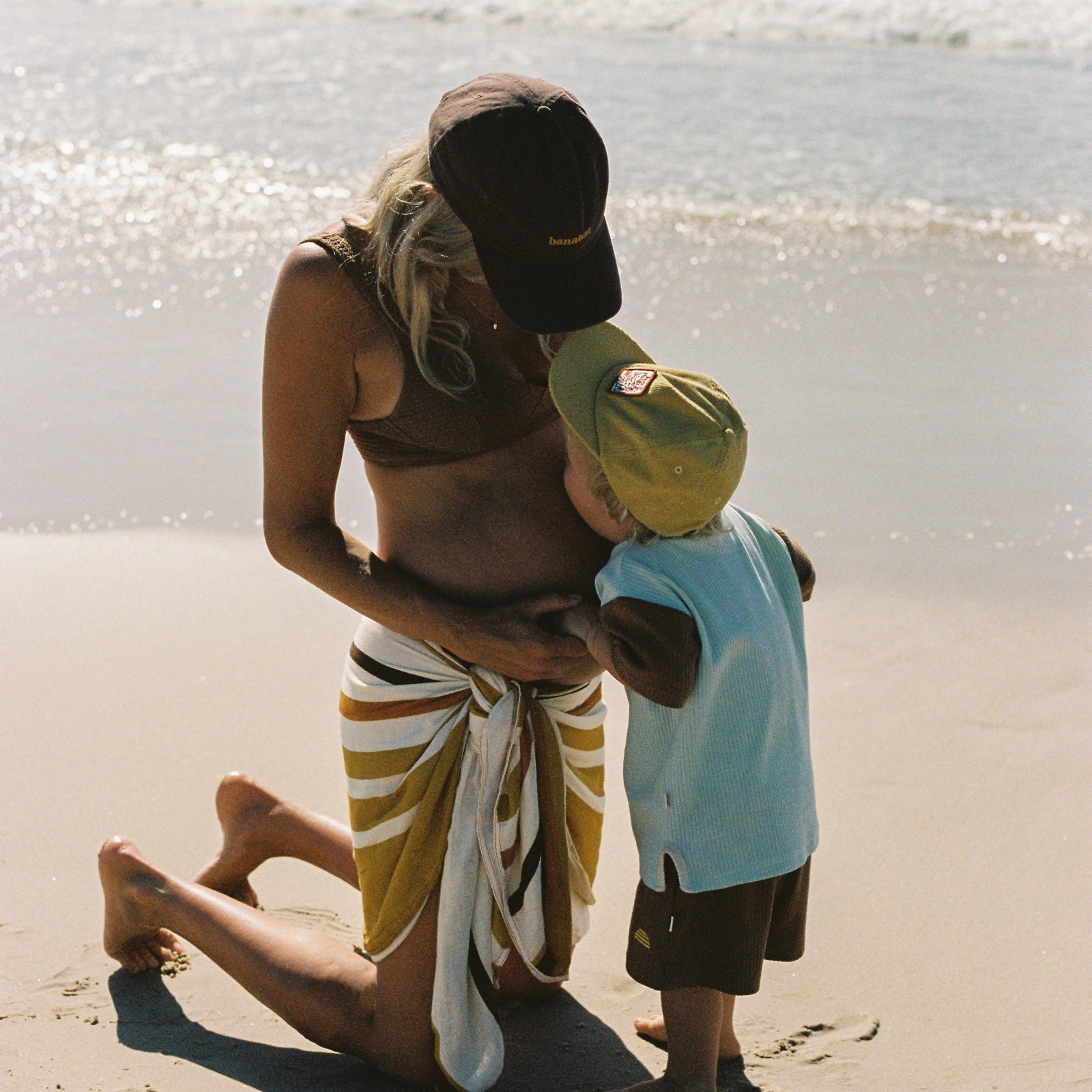 lifestyle image of pregnant women on the beach wearing a  brown bikini top, Banabae cap in brown with Banabae embroidered on the center front in yellow and Banabae Golden Daze Sarong tied around waist. Looking down at a small child wearing Banabae Park Ha